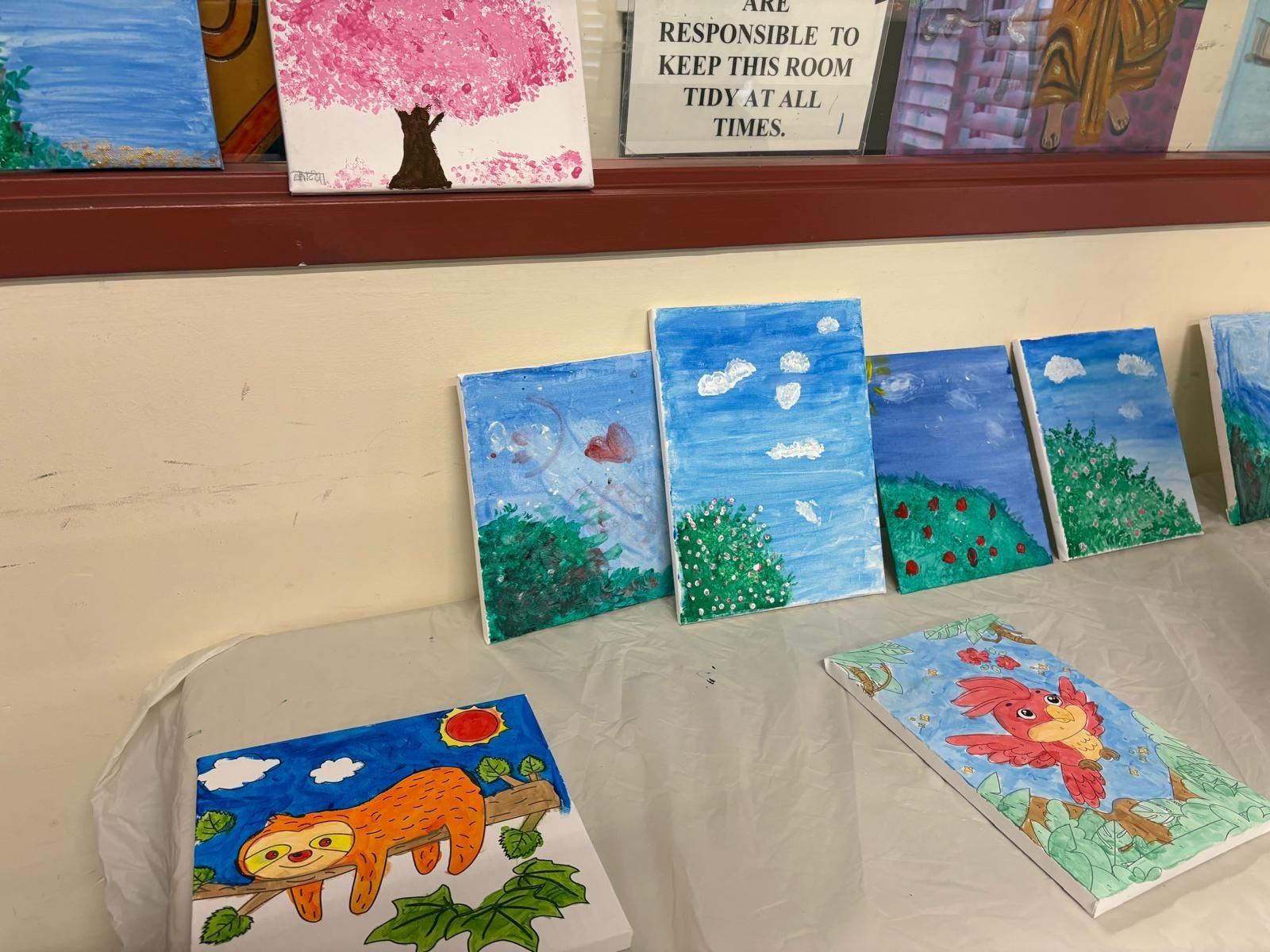 Series of colorful paintings on a table, featuring nature scenes and animals, with a poster above.