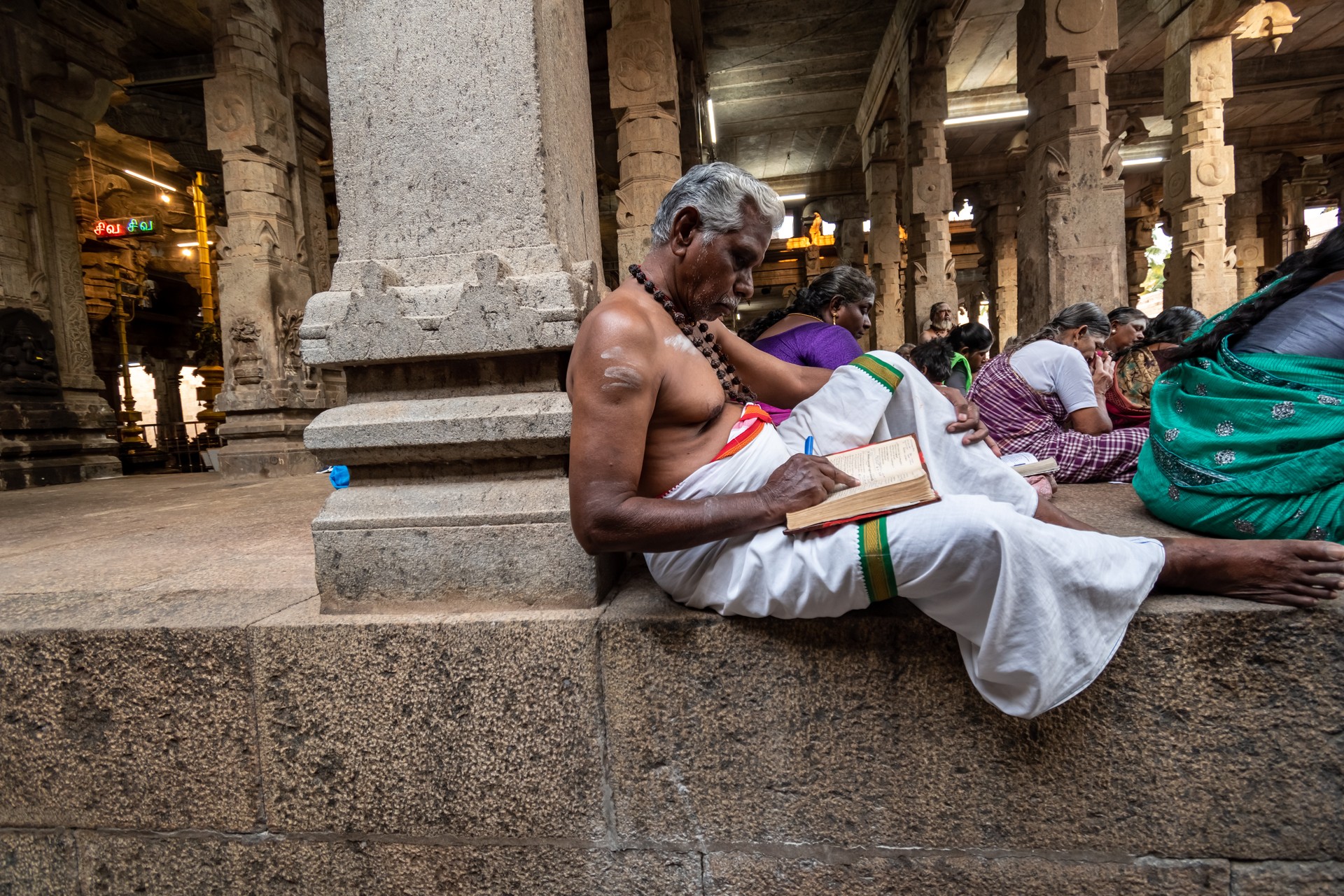 An elderly Hindu temple priest in traditional attire reading a book